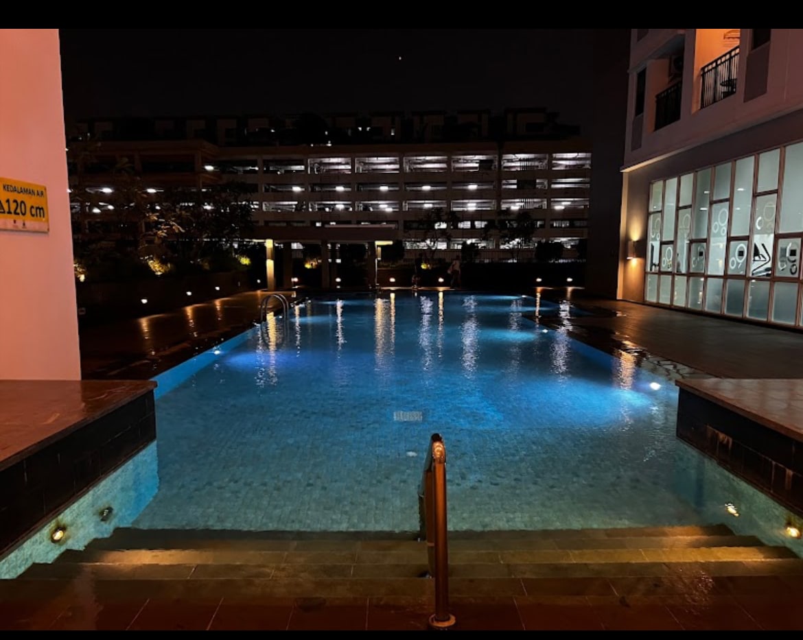 The outdoor pool is illuminated with soft blue lighting, creating a calm ambiance at night. Surrounding greenery and a spacious deck complement the area. In the background, the parking structure is subtly visible, enhancing the sense of safety and convenience.