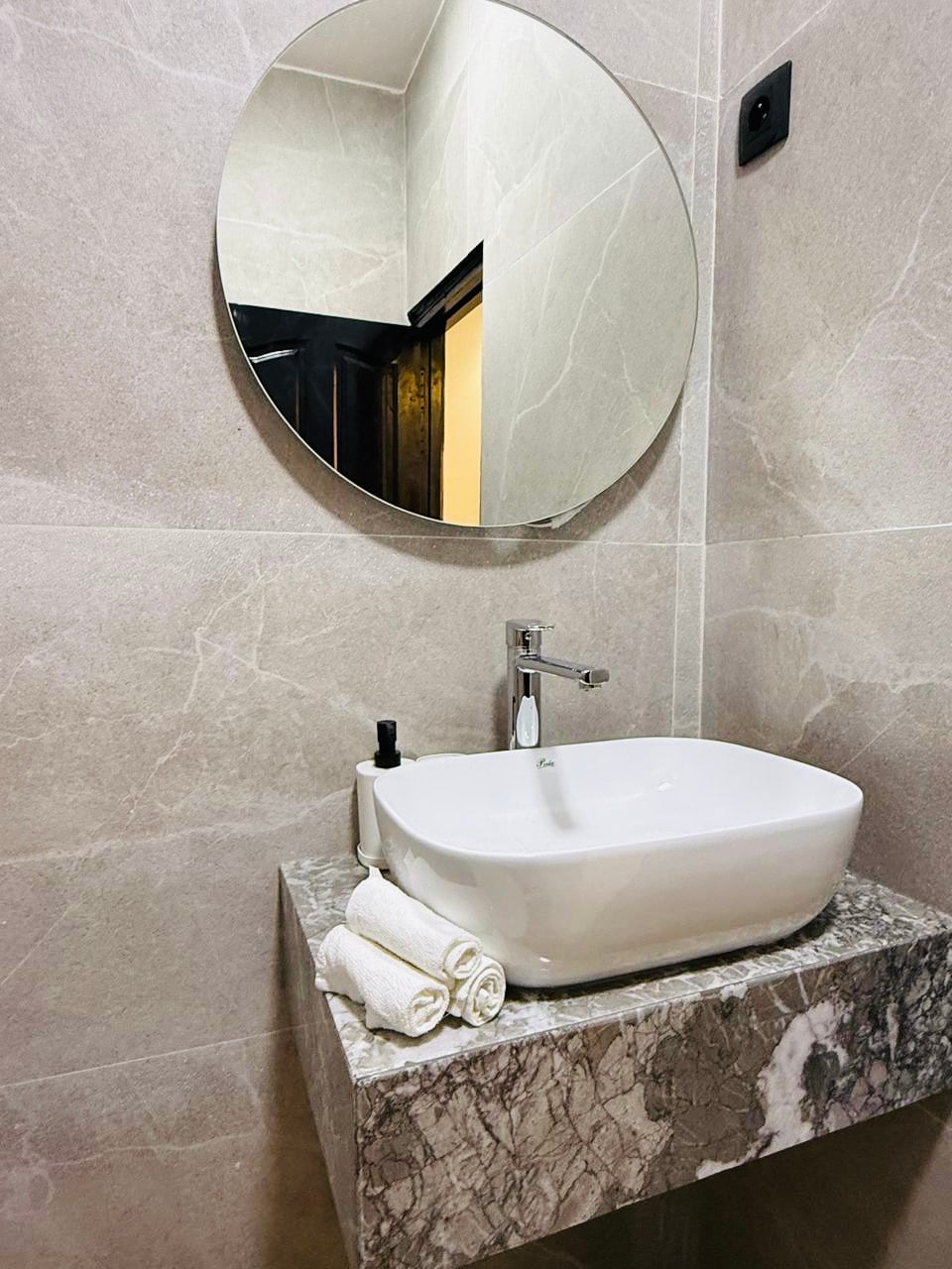 A modern restroom is showcased, featuring a stylish wall-mounted sink atop a marble countertop. A round mirror reflects the space, and neatly rolled towels are placed beside the sink. The soft, neutral tones of the wall tiles create a clean and sophisticated ambience.