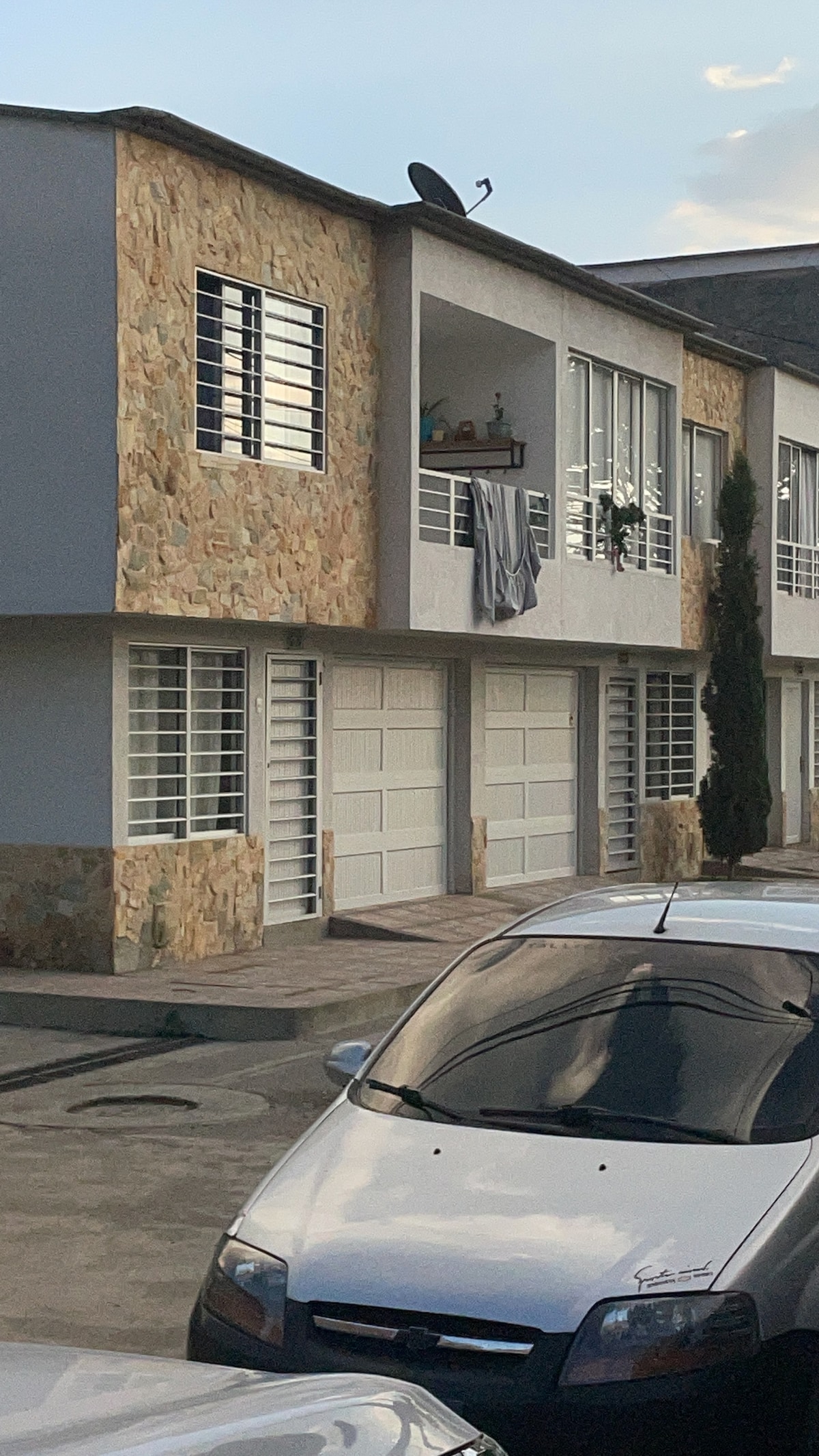 A two-story building is visible, featuring a combination of stone and smooth white exterior. Several large windows are arranged symmetrically on the facade. A gray car is parked in the foreground, and a balcony with personal items is seen on the upper level.