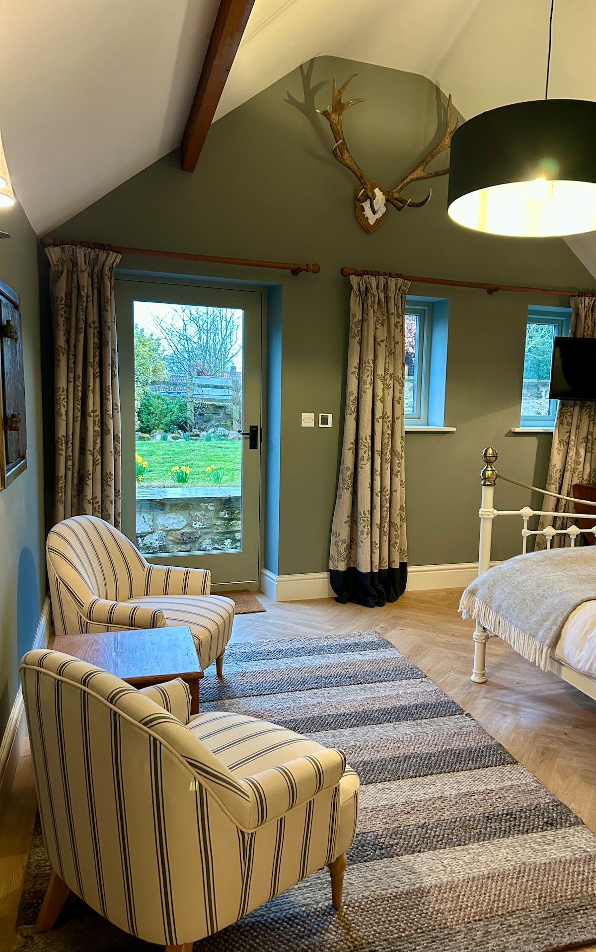 A bright room features two striped armchairs facing a door that opens to a green outdoor space. Natural light streams in through large windows adorned with patterned curtains. A soft area rug complements the wooden flooring, adding a touch of warmth to the atmosphere.