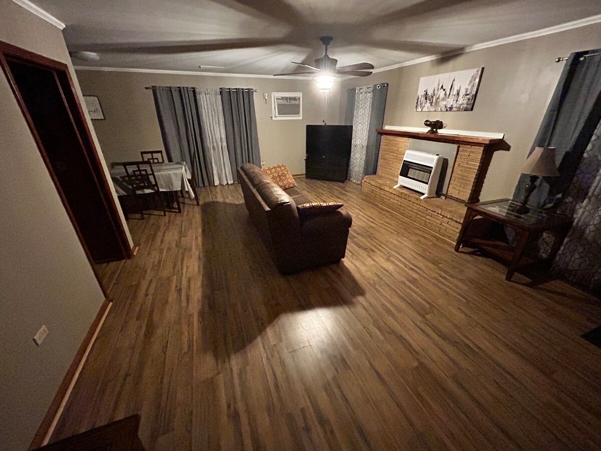 A spacious living area features a comfortable brown sofa facing a large television. A wooden coffee table is positioned in front of the sofa, while a dining table with chairs is visible in the corner. Light wood flooring is complemented by grey curtains covering the windows.