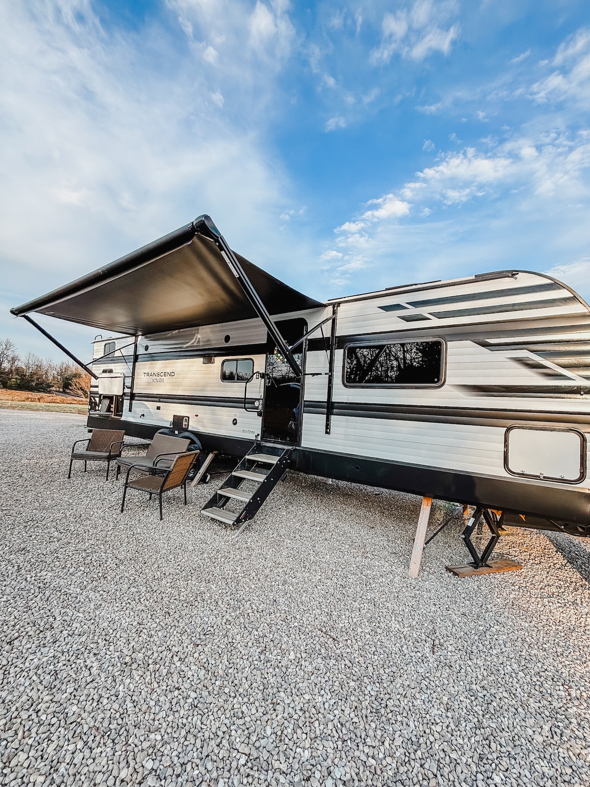 The exterior of the RV is shown, featuring a large awning that provides shade for outdoor seating. Two chairs are positioned on gravel, and a set of stairs leads into the RV. The sky above is clear with scattered clouds.
