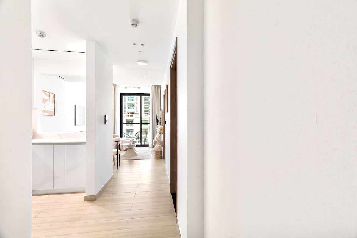 A bright corridor leads towards an open living space and a private balcony. Smooth, light-colored tiles add warmth, while natural light filters through large windows. Minimalist decor enhances the clean lines, and a cozy seating area can be glimpsed through the doorway.