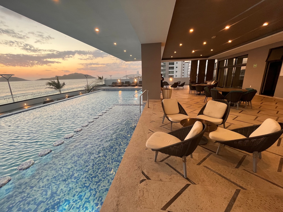 An outdoor pool area is visible with a tranquil surface reflecting the sunset. Comfortable seating is arranged nearby, featuring modern chairs and tables. The shoreline is framed in the background, highlighting the spacious atmosphere and scenic views.