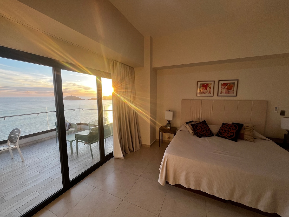 A spacious bedroom is designed with large glass doors that open to a stunning ocean view. Soft sunlight enters the room, highlighting the inviting bed and area features. A seating area is visible on the balcony, providing an ideal spot for relaxation with scenic vistas.