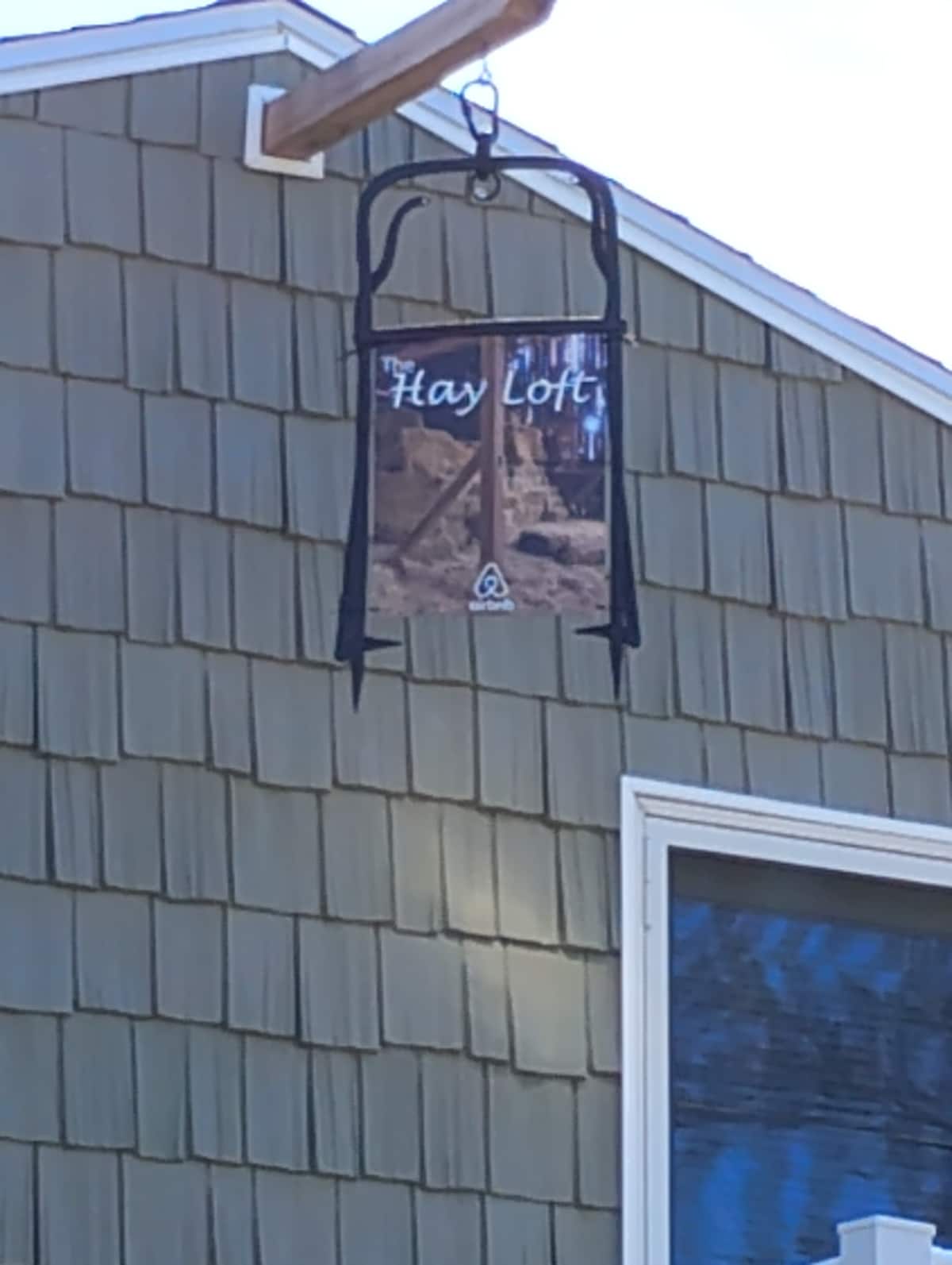 A sign displaying 'Hay Loft' is prominently hung from a wooden fixture on a shingled exterior wall. The sign features an image of trees and natural scenery, indicating a welcoming entrance to the property. Sunlight reflects off a nearby window.