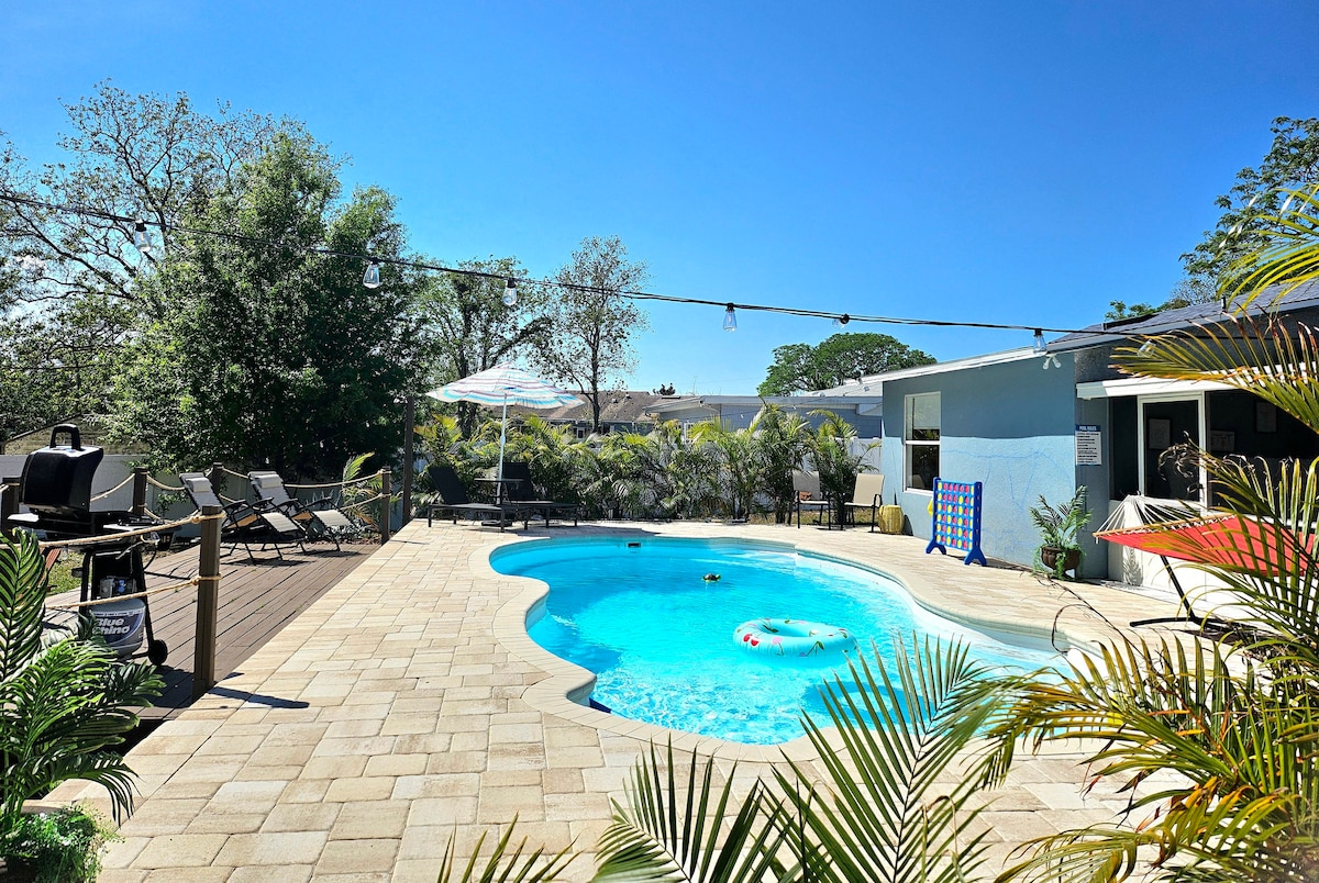 A private pool is surrounded by stone tiles and lush greenery, providing a relaxing outdoor space. Sun loungers are arranged along the poolside, while an umbrella offers shade. A barbecue grill is visible nearby, and a game board can be seen against the wall.