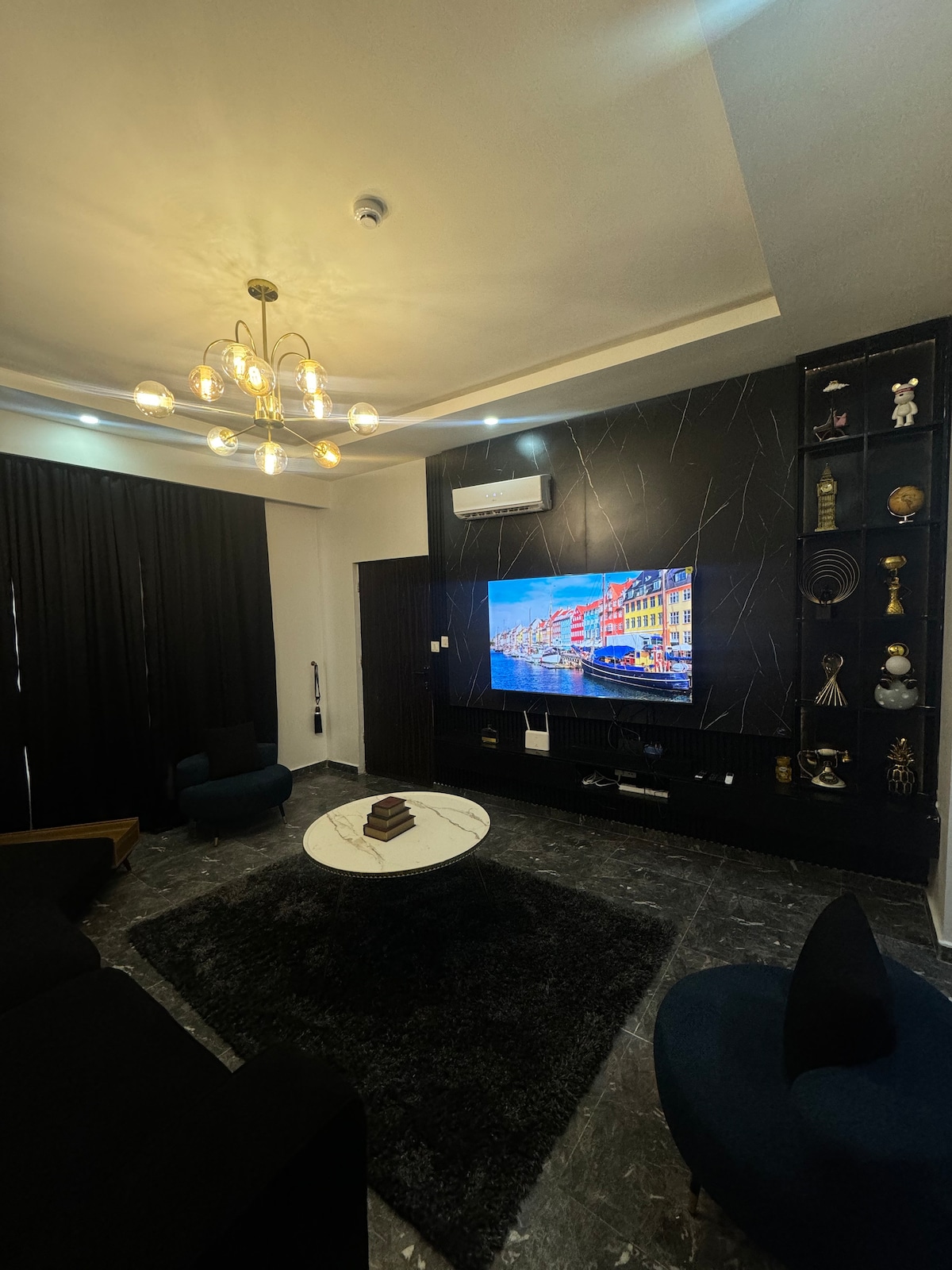 A modern living area features a sleek black accent wall with a large flat-screen television. A marble coffee table is situated on a plush dark rug. Two stylish chairs in contrasting blue are arranged alongside a comfortable black sofa. Elegant lighting fixtures enhance the ambiance.