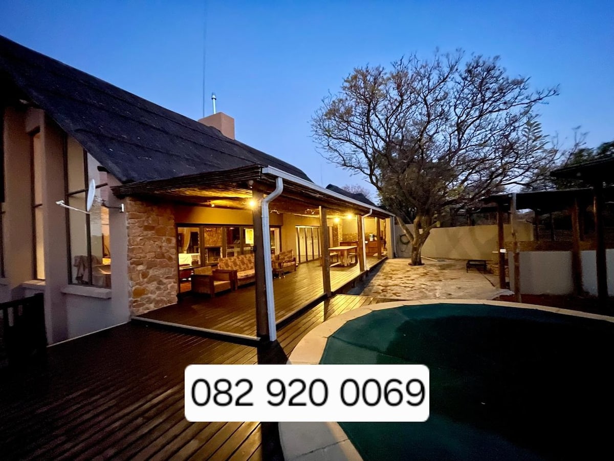 An outdoor view of the house at dusk, highlighting the wooden deck and cozy seating area. Soft lighting illuminates the porch, creating a welcoming ambiance. A large tree stands nearby, and a pool is visible in the foreground, surrounded by a stone patio.