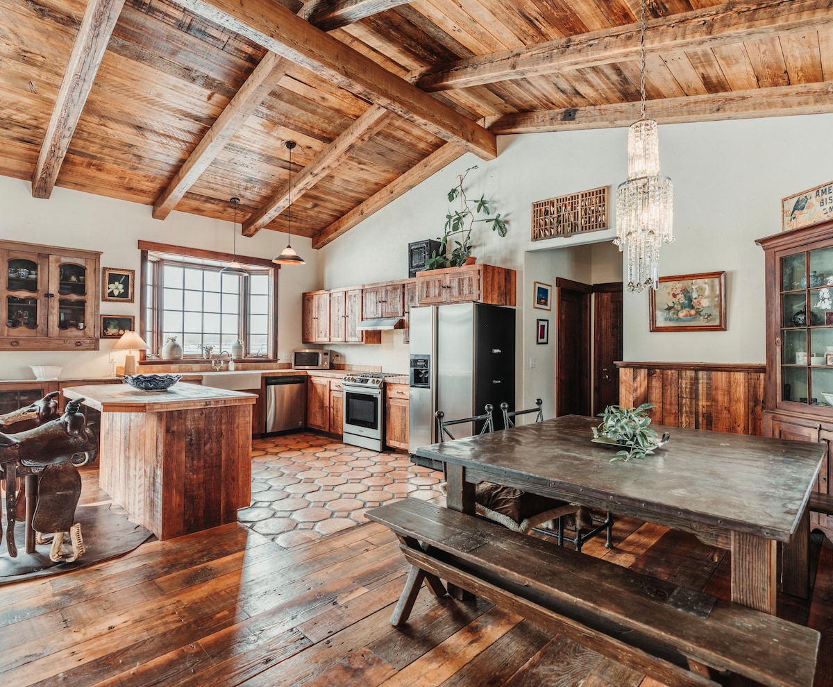 A spacious kitchen and dining area feature high vaulted ceilings with exposed wooden beams. Sterling appliances line the countertops, complemented by wooden cabinets. A large wooden dining table with bench seating invites gatherings, while warm tones and rustic details enhance the overall ambiance.