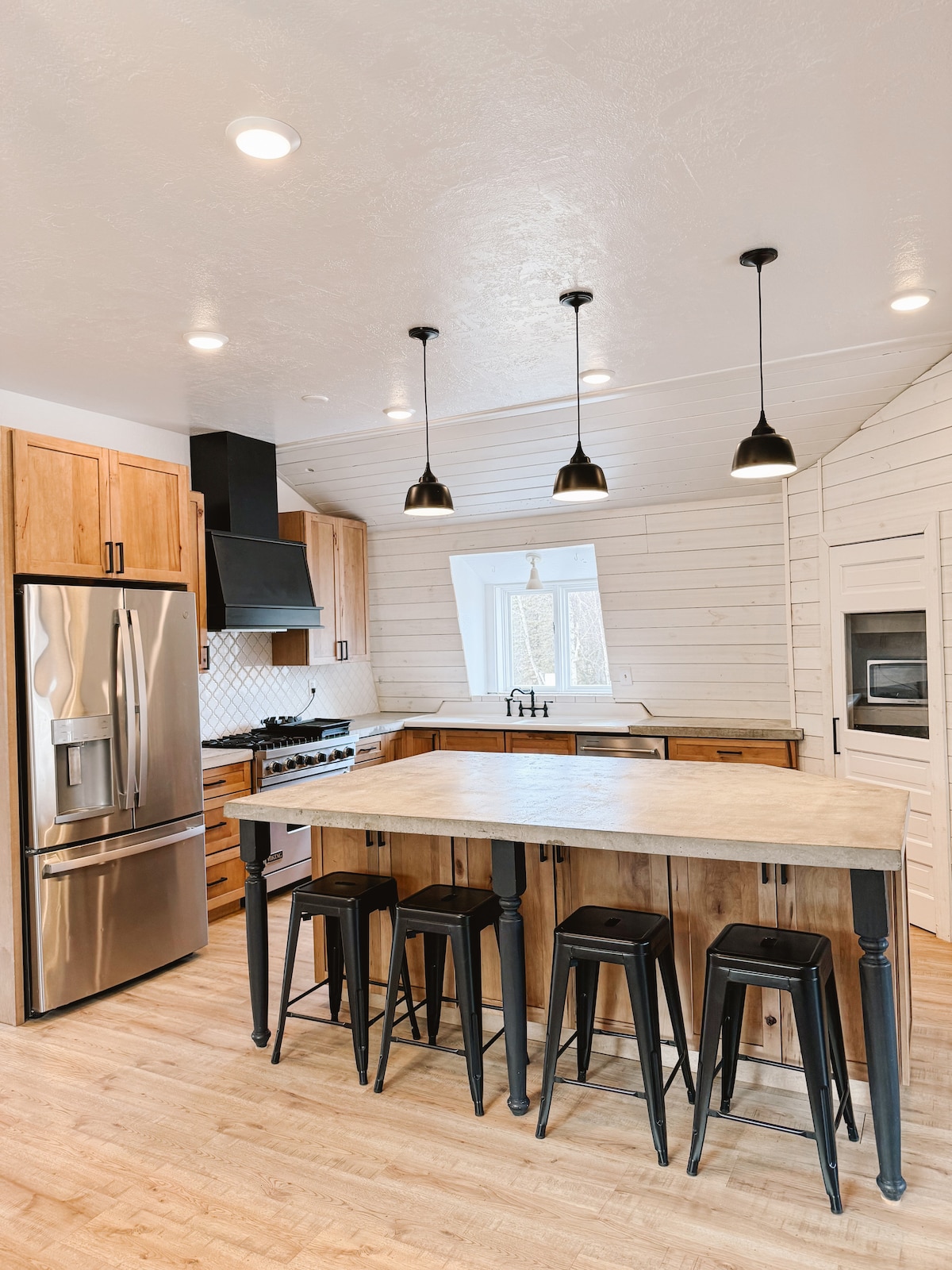 A modern kitchen is presented with light wood cabinetry and stainless steel appliances. A central island with seating for four is complemented by pendant lighting. Natural light enters through a nearby window, enhancing the clean design of the space.