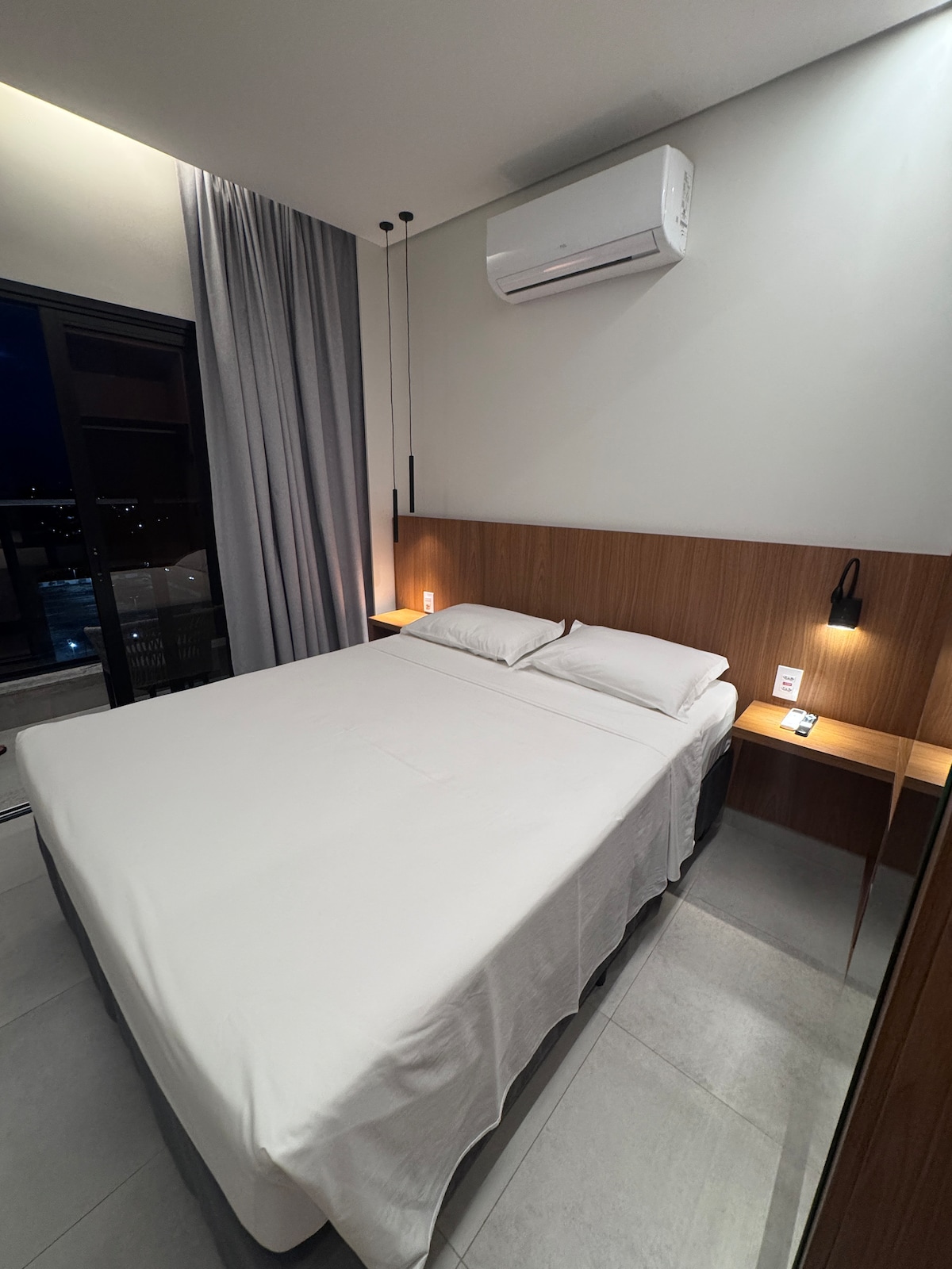 A modern bedroom features a spacious bed neatly dressed in white linens. Soft lighting is provided by wall-mounted fixtures on either side of the bed. A large window with light curtains offers a view of the outside, while gray curtains frame the window.
