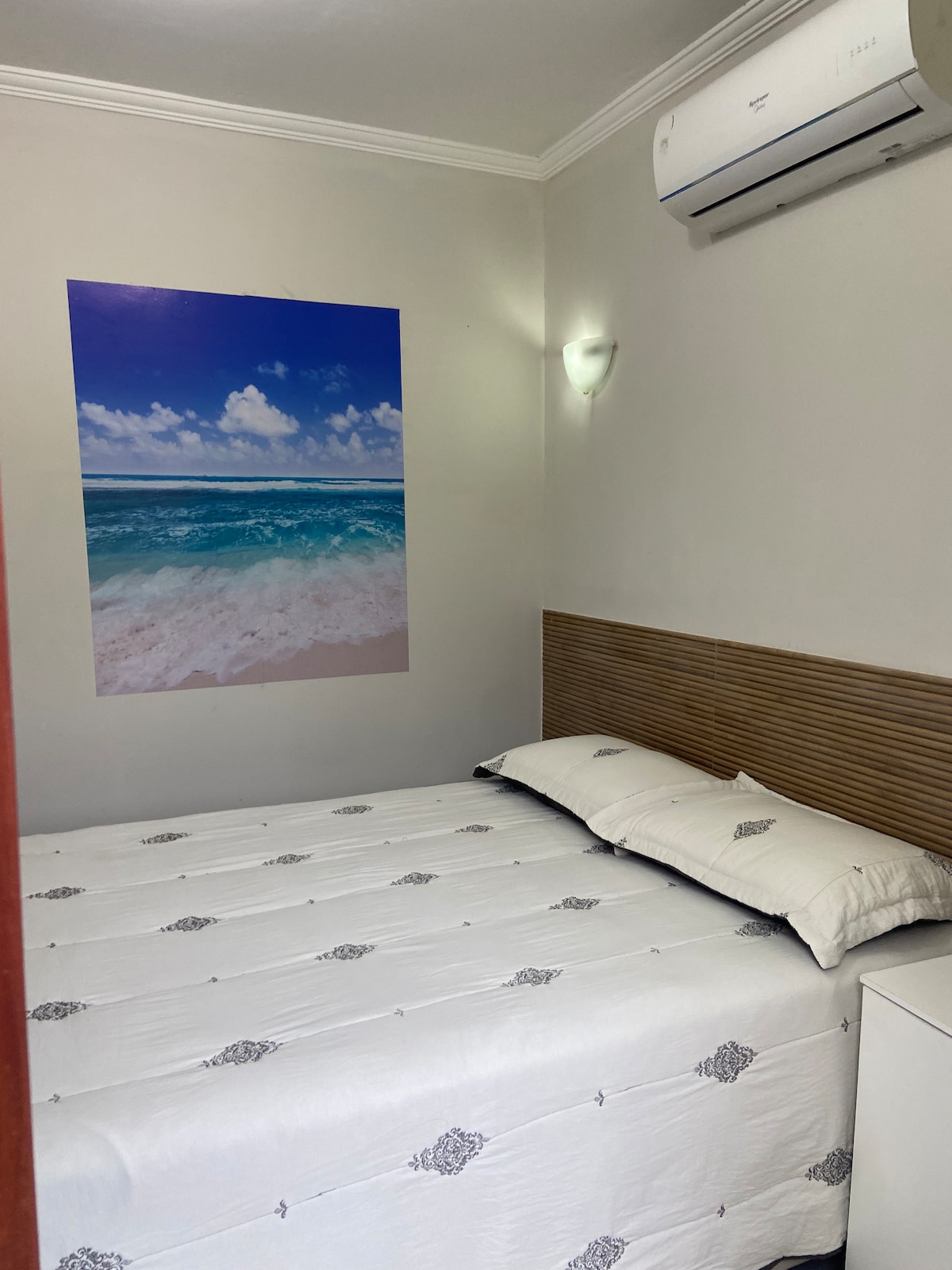 A compact suite is shown, featuring a spacious bed with a white coverlet and decorative black patterns. A calming beach mural adorns one wall, bringing a sense of tranquility. An air conditioning unit is installed overhead, and a bedside table is positioned nearby.