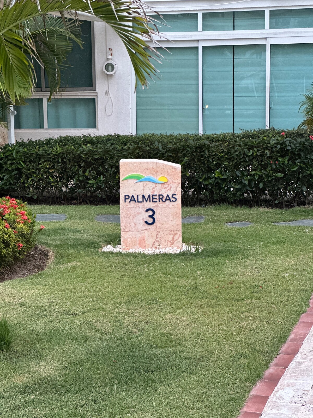 A stone sign displays the number '3' and the name 'PALMERAS' in blue and yellow lettering. The sign is surrounded by neatly manicured grass and small white flowers, with green shrubbery and palm leaves visible in the background.