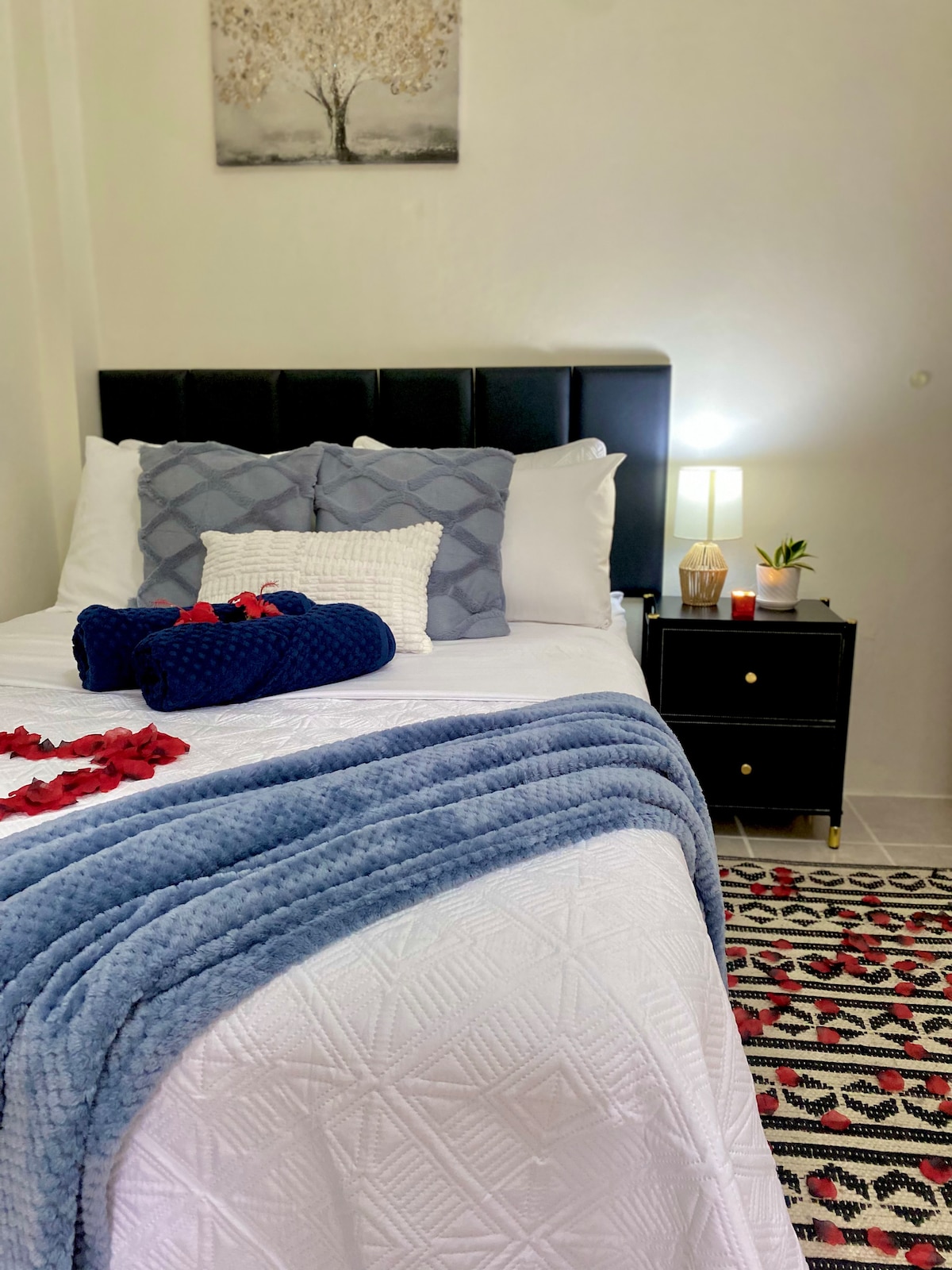 A cozy sleeping area features a spacious bed adorned with a white quilt and decorative pillows. A bedside table holds a lamp and small plants. A patterned rug adds warmth to the space, while a serene piece of wall art complements the decor.