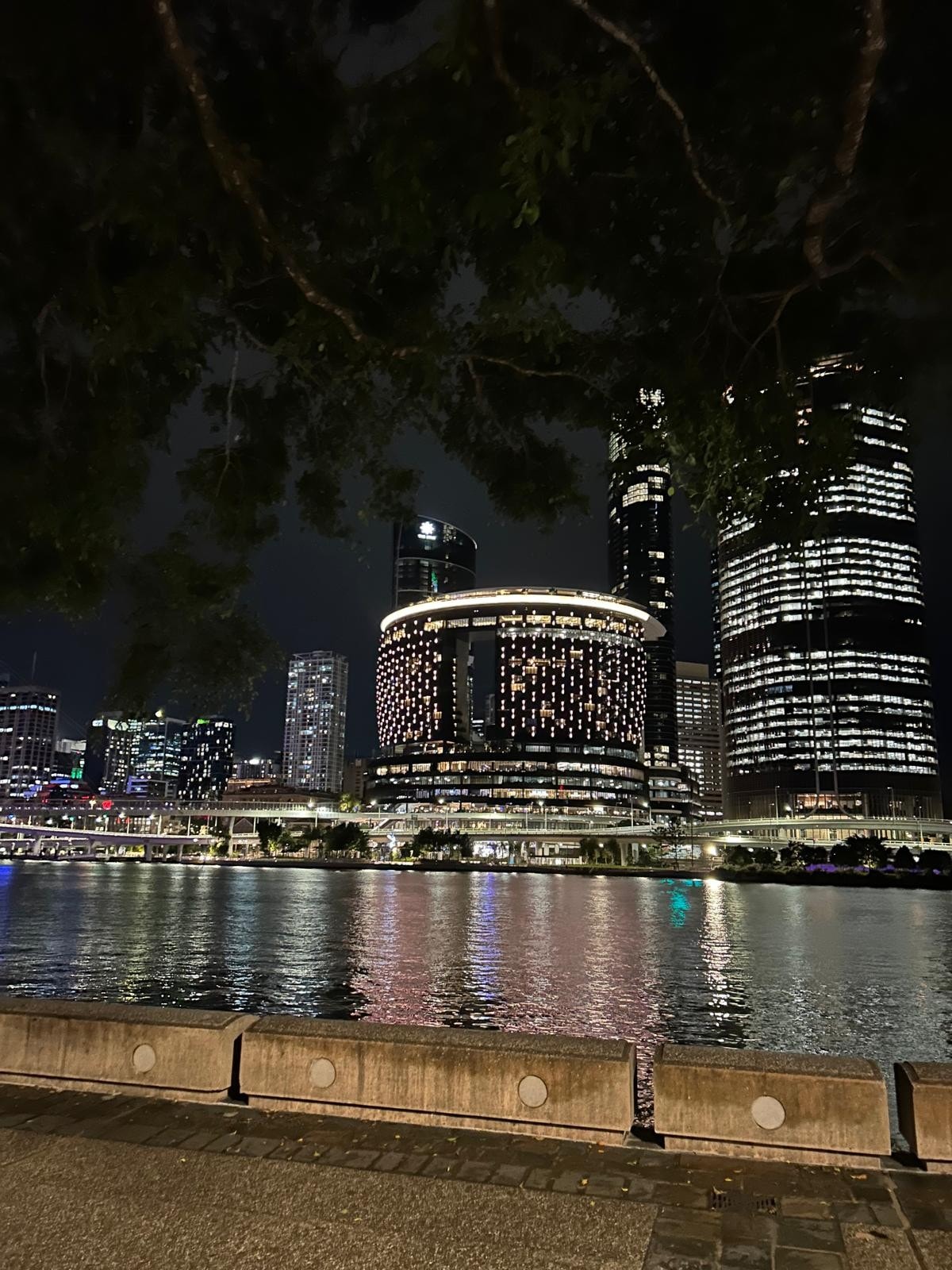 A serene nighttime view features modern high-rise buildings reflecting in the calm water. The prominent circular structure is adorned with bright lights, creating a vibrant atmosphere along the waterfront. Trees provide a natural frame, enhancing the tranquil urban scene.