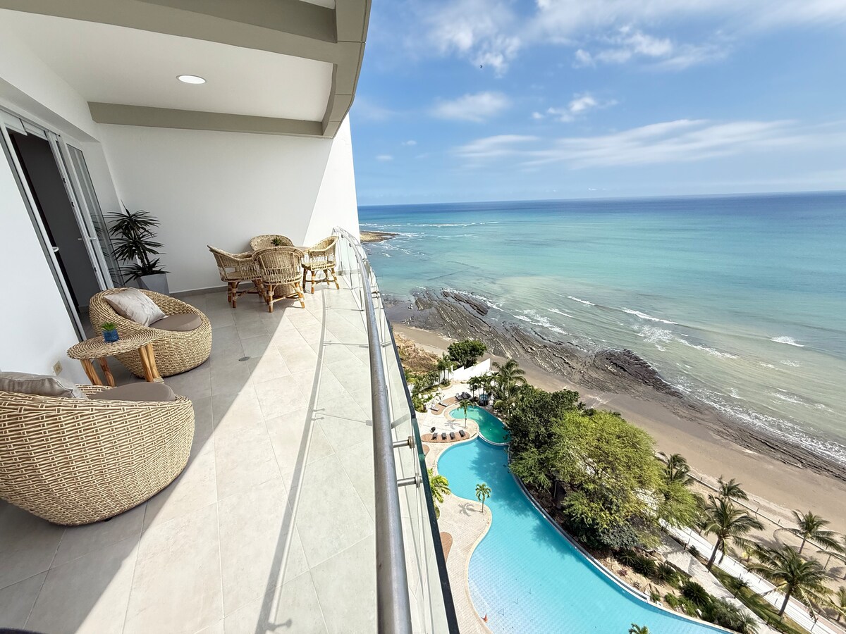 A spacious balcony offers serene views of the ocean and coastline. Two comfortable wicker chairs are positioned for relaxation, accompanied by a small table. A sparkling pool is visible below, surrounded by palm trees and sandy beach, enhancing the inviting outdoor space.