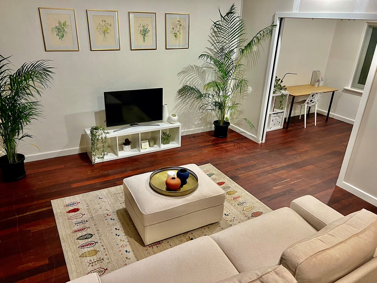 A cozy living area is highlighted by a light-colored sectional sofa facing a modern television. Decorative plants add a touch of greenery, while framed botanical art complements the neutral walls. A stylish coffee table sits in the center, enhancing the spacious ambiance.
