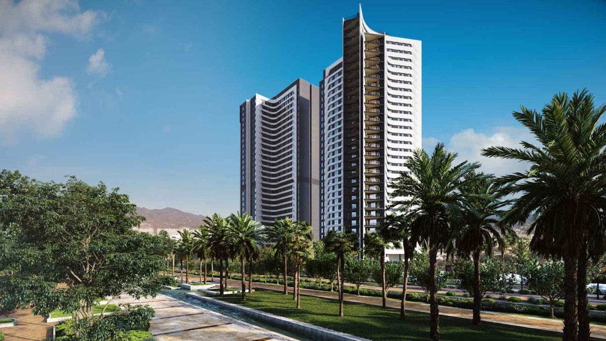 Two tall residential buildings are visible against a clear blue sky, showcasing modern architectural designs. Lush palm trees and manicured greenery line a pathway in the foreground, creating a serene setting. The background reveals gentle mountains, adding depth to the landscape.