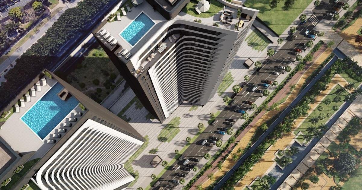Aerial view of two modern towers, showcasing their unique architectural design. Rooftop pools can be seen on several floors, accompanied by landscaped areas. Surrounding the buildings, a parking area is visible, along with well-maintained pathways and greenery, indicating a tranquil environment.