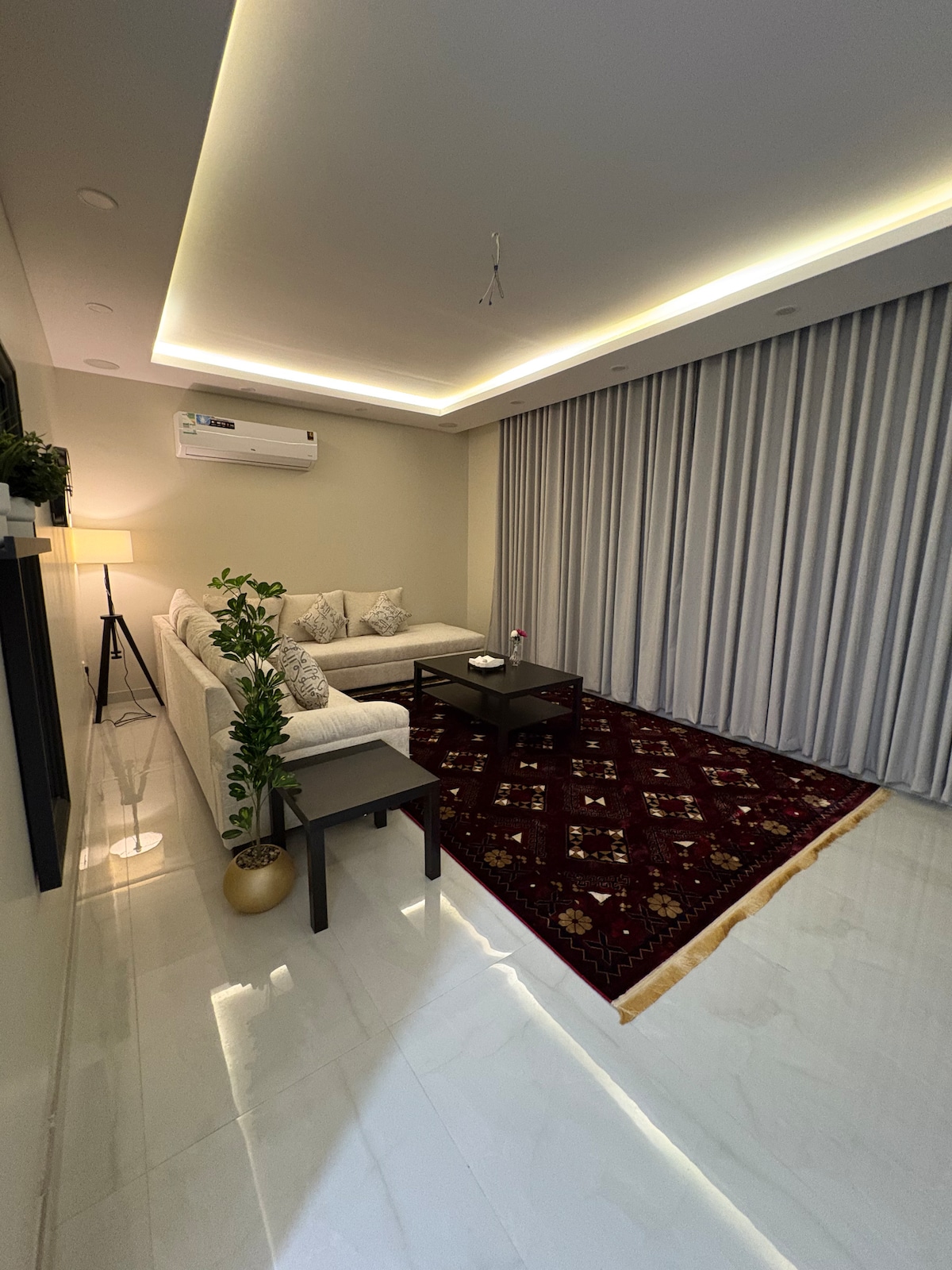 A modern living area features a light-colored sectional sofa decorated with patterned cushions. A dark coffee table sits centrally on a richly colored rug. Soft lighting highlights the minimalist decor, while floor-to-ceiling curtains cover a large window, enhancing the serene ambiance.