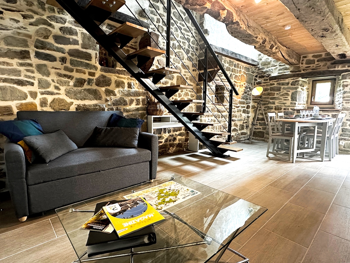 An interior space featuring exposed stone walls and wooden beams. A comfortable gray sofa is positioned next to a glass coffee table adorned with a magazine. A dining table with chairs is visible, complemented by a staircase leading to an upper level, creating an inviting common area.