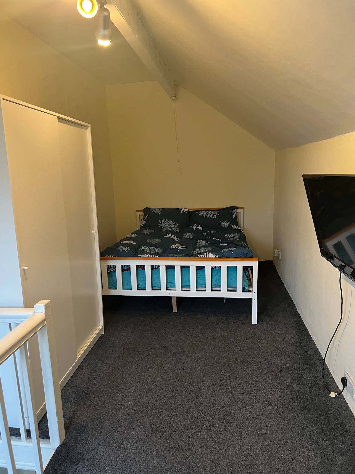 A cozy sleeping area is highlighted by a compact bed dressed in a blue and white patterned duvet. A wardrobe stands alongside the bed, while a television is mounted on the wall opposite. Soft carpeting covers the floor, and angled ceilings create an intimate space.