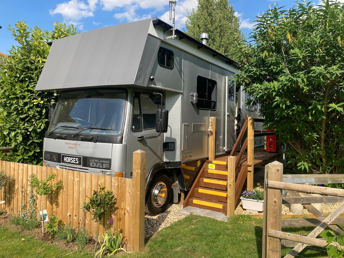 A horse lorry is parked beside a wooden fence, with steps leading to the entrance. The vehicle's silver exterior reflects sunlight, while lush greenery surrounds the area. A gravel path is visible, offering access through a wooden gate, providing a sense of privacy and tranquility.
