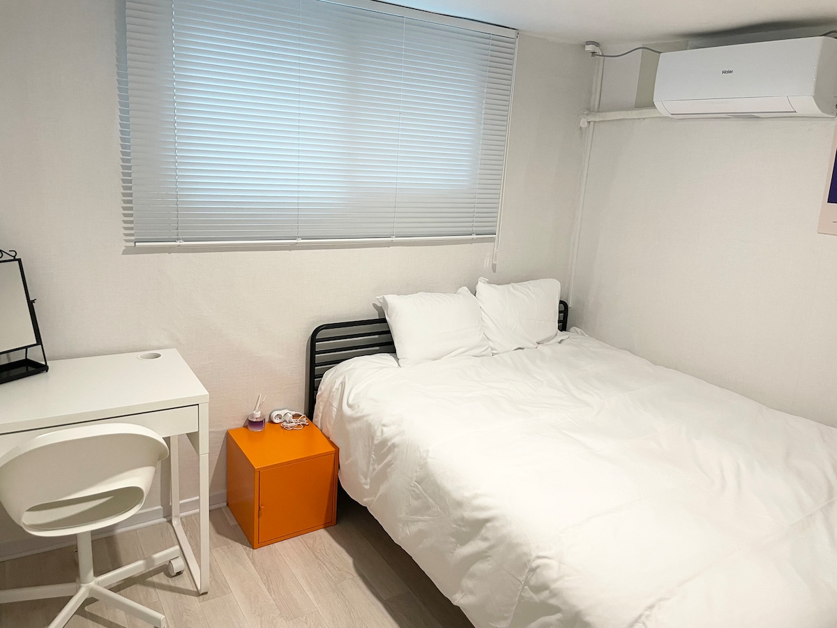 A neatly arranged sleeping area features a spacious bed dressed in white bedding, complemented by white pillows. An orange nightstand stands beside the bed, contrasting gently with the neutral-colored walls. A minimalist desk and chair are positioned against one wall, beneath a window with light-filtering blinds.