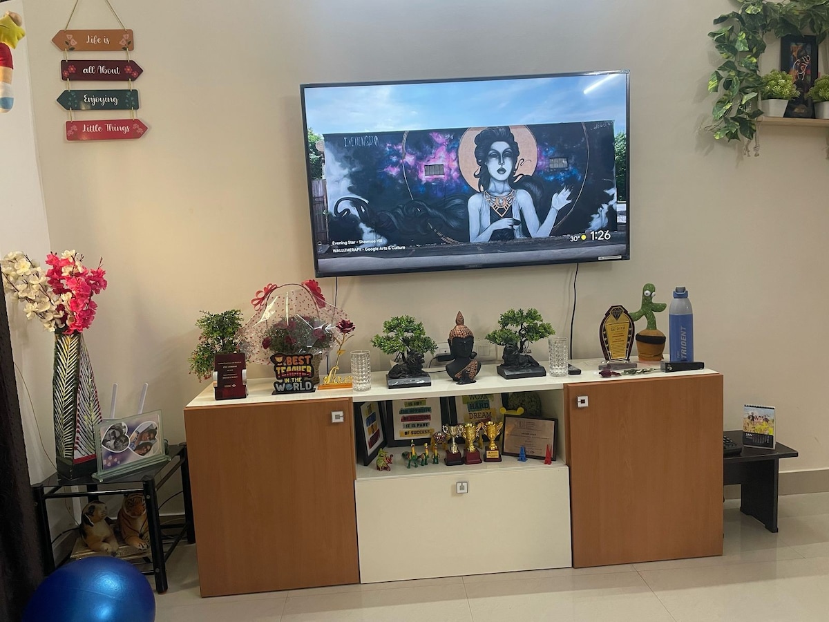 A media console is positioned against a wall, displaying a large television. Various decorative items, including plants and figurines, are arranged on top. A wall shelf holds additional decor, while colorful art is visible on the screen, contributing to a vibrant living area.