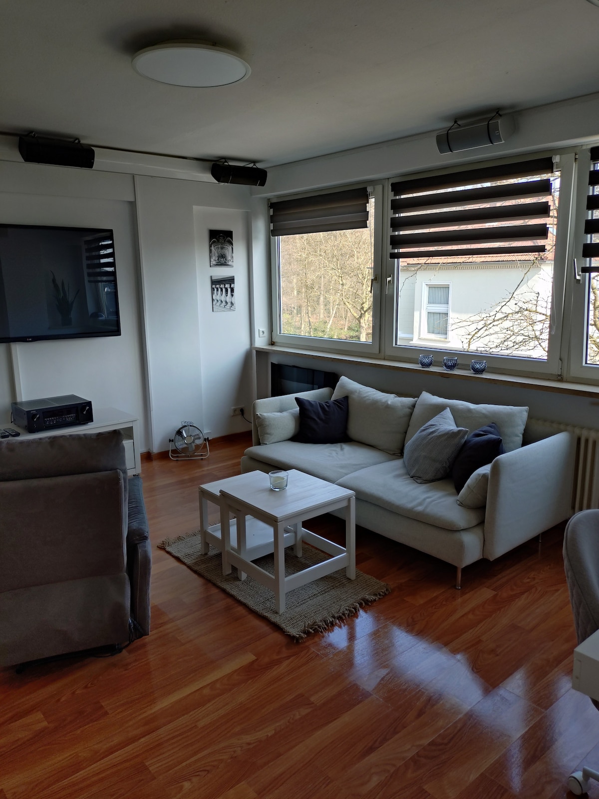 A modern living area features large windows with horizontal blinds, allowing natural light to fill the space. A light-colored sofa is arranged with decorative pillows, accompanied by a small coffee table atop a woven rug. A flat-screen TV is mounted on the wall, enhancing the entertainment options.