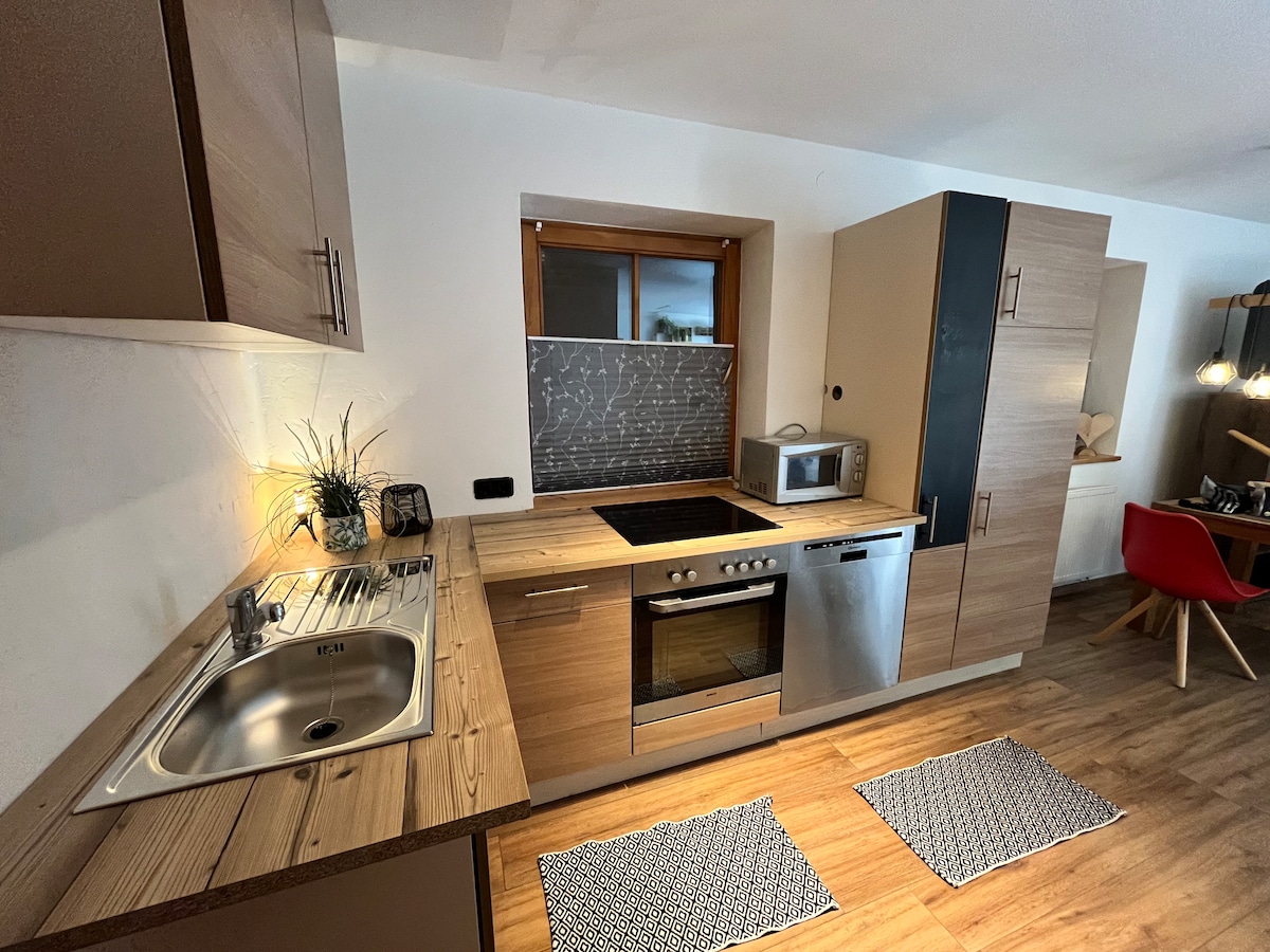 A modern kitchen is displayed with sleek wooden cabinetry and a coordinated countertop. An integrated cooktop and oven are visible, alongside a microwave and a sink. Two patterned mats lay on the floor, contributing to a functional cooking environment. Natural light filters through a nearby window.