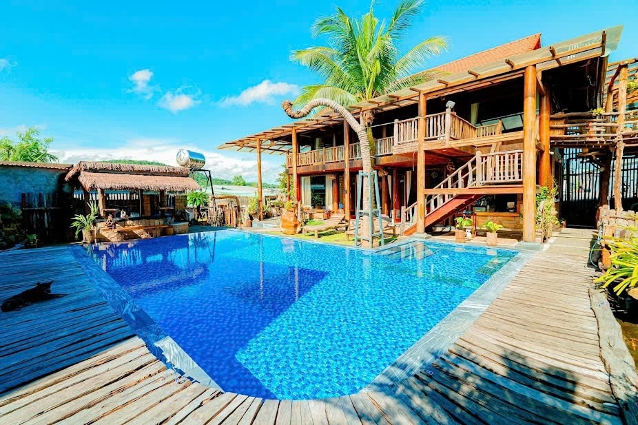 An expansive outdoor area features a large swimming pool surrounded by a wooden deck. The building, made of natural materials, provides multiple levels with balconies and staircases visible. Lush greenery complements the setting, while palm trees sway gently in the background.