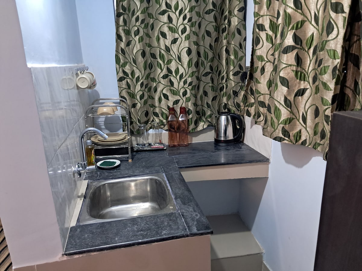 A compact kitchenette area is visible, featuring a stainless steel sink and a dark countertop. Modern appliances, including an electric kettle and several glass bottles, are arranged neatly. Light filters through patterned curtains, adding a touch of warmth to the space.