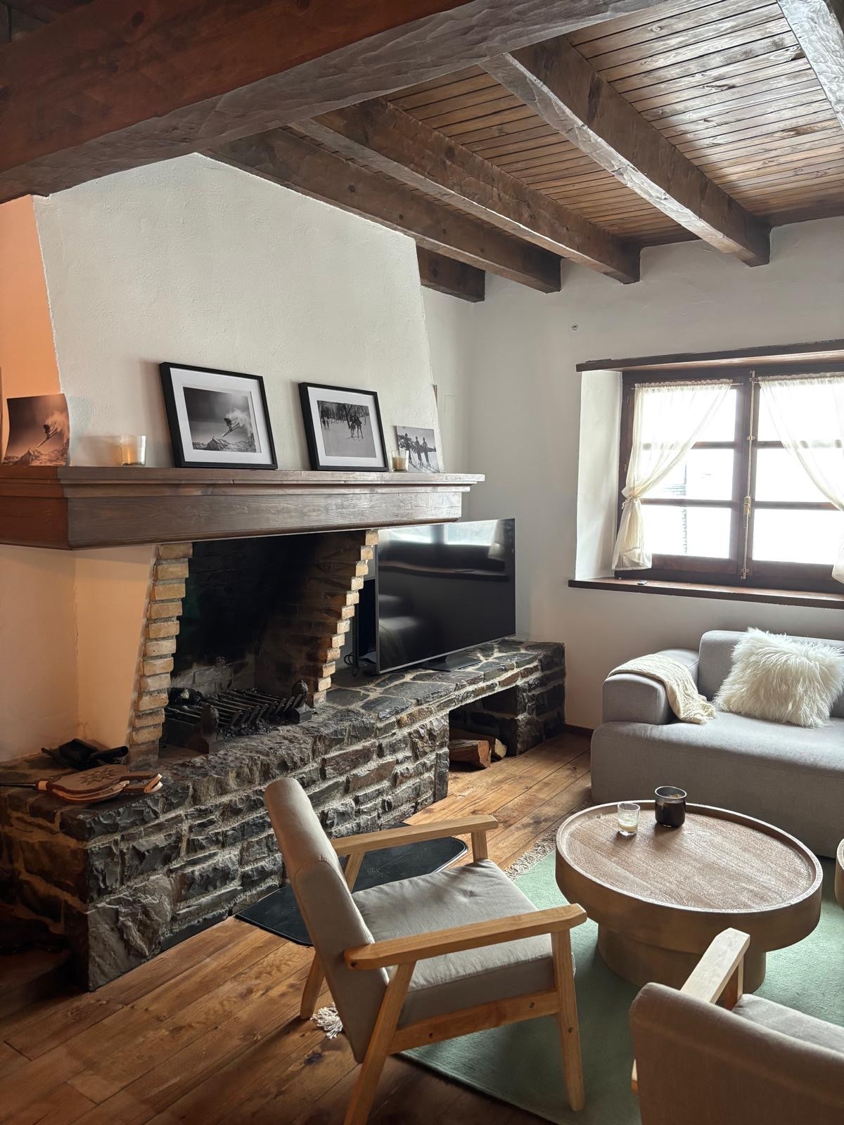 A rustic living area features a stone fireplace with wooden mantel, framed black-and-white photographs above. A soft gray sofa and a circular wood coffee table are complemented by a light rug. Natural light is filtered through a window dressed in sheer curtains.