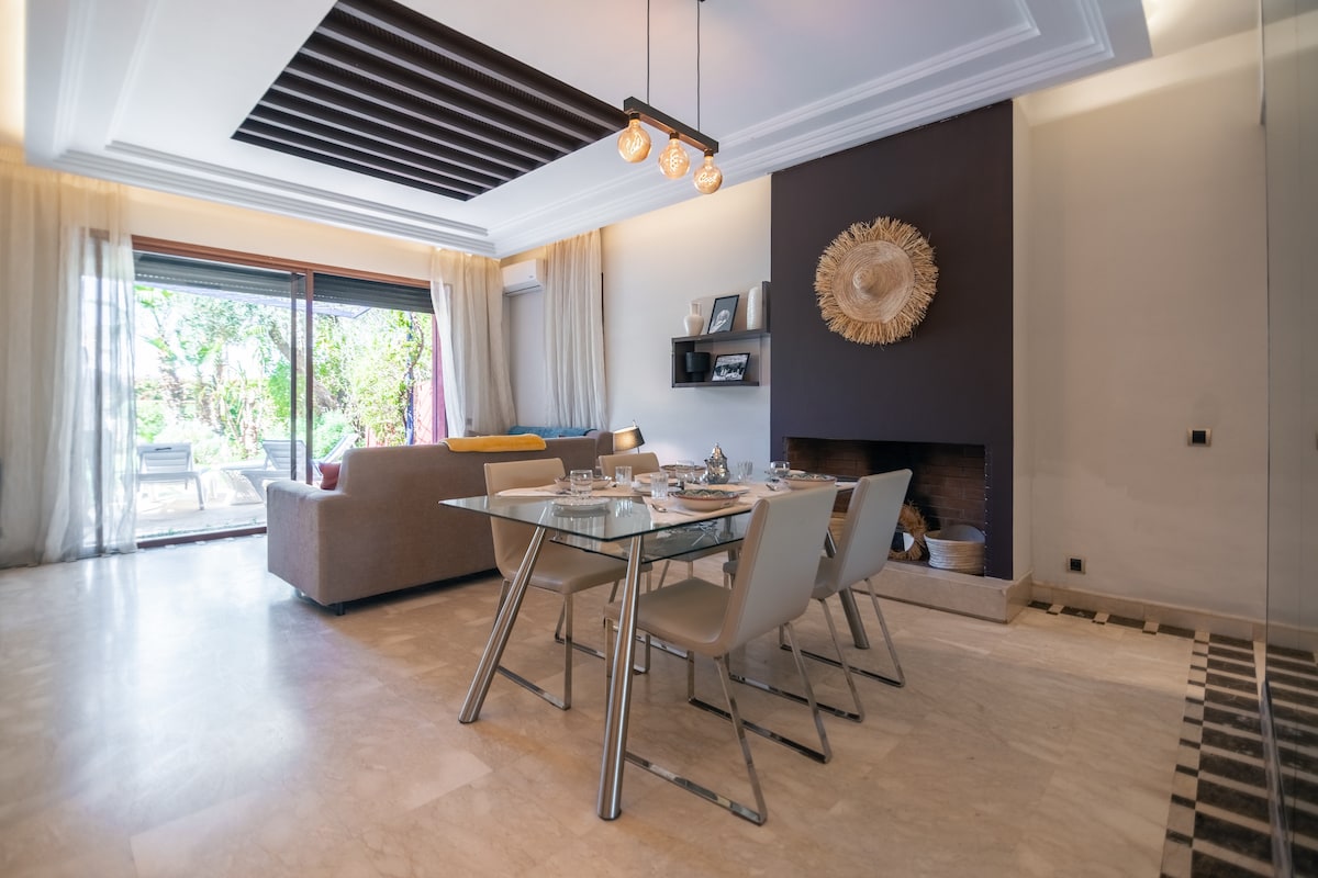 A modern dining area is showcased, featuring a glass dining table set with elegant dinnerware. Comfortable chairs are arranged around the table, with soft lighting provided by overhead fixtures. A spacious living area is visible, leading to a terrace with outdoor seating and garden views.