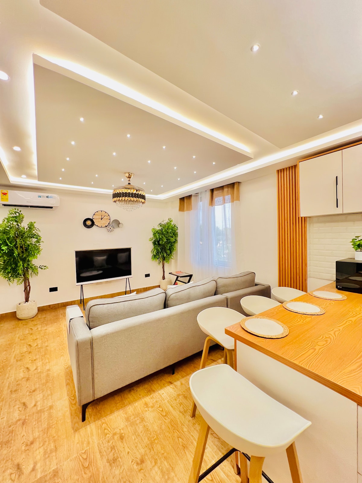The spacious living area showcases a modern design with a comfortable sofa facing a flat-screen TV. Natural light filters through large windows, and artificial lighting highlights the elegant ceiling. Potted plants add a touch of greenery, while the open kitchen features bar seating with minimalist stools.