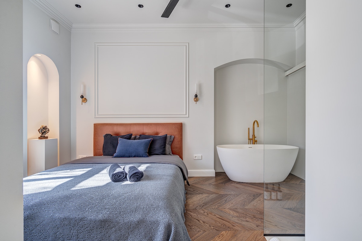 A serene bedroom features a spacious bed with muted linens and decorative pillows, complemented by a modern freestanding bathtub nearby. Soft lighting is provided by wall sconces, and warm wood flooring adds texture to the inviting space.
