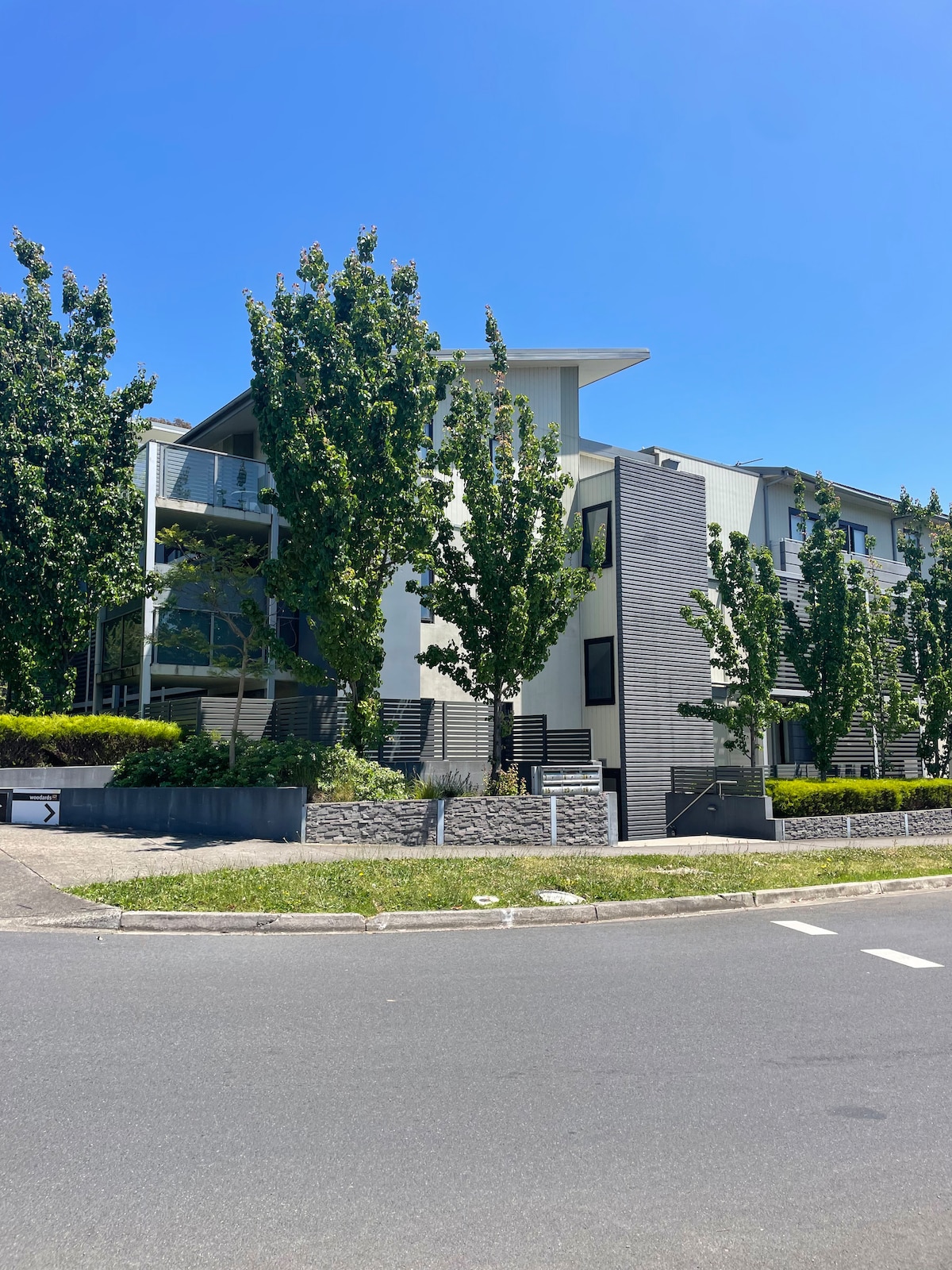 The modern apartment building is framed by lush, tall trees against a clear blue sky. The structure features a combination of stone and sleek façade elements, with several balconies visible on the upper floors. A manicured green space is included at the base.