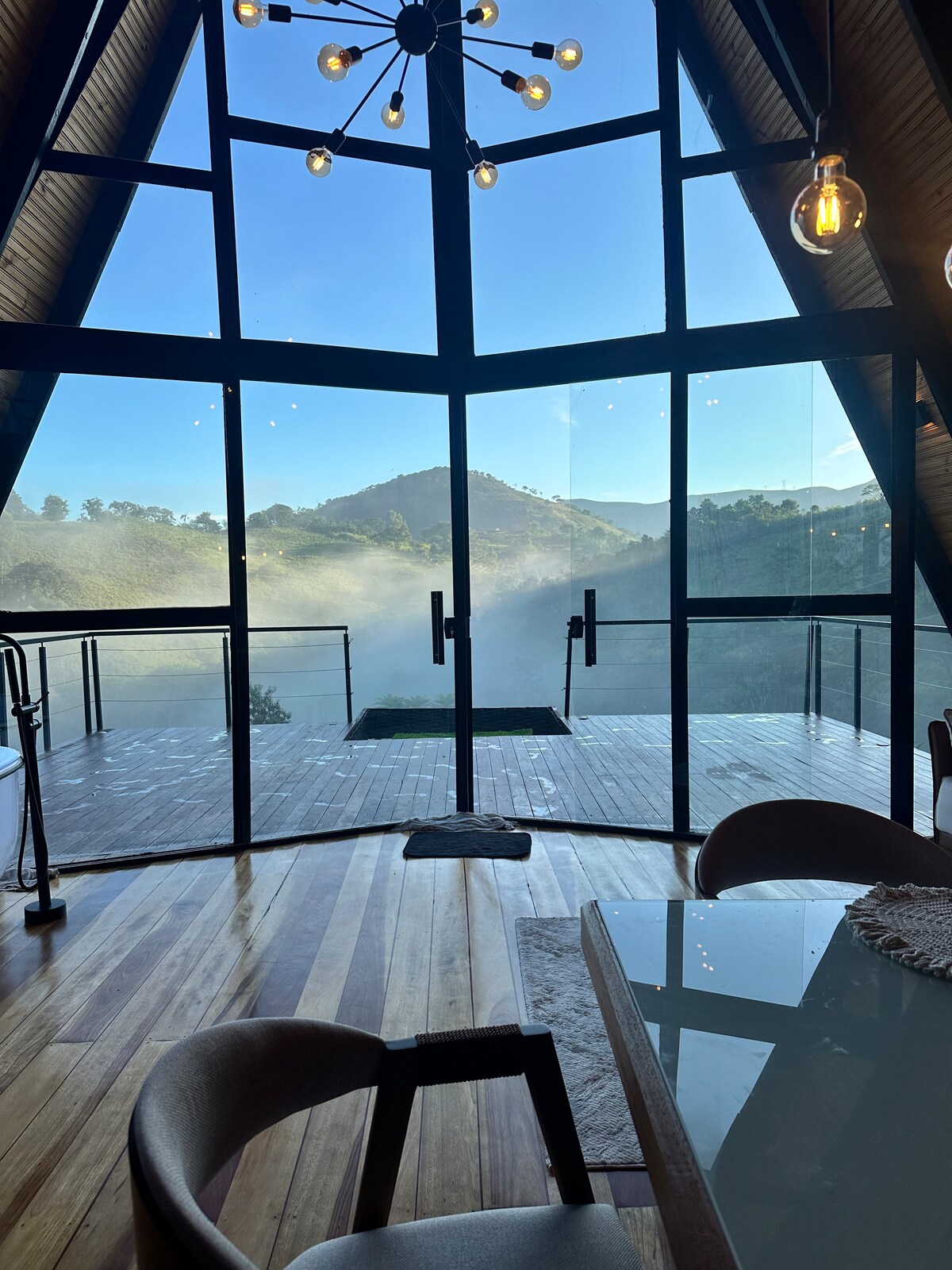 A large glass wall allows for unobstructed views of the surrounding hills, with mist softly enveloping the landscape. Natural light fills the space, where wooden flooring and modern furnishings create an inviting interior. A contemporary light fixture adds a stylish touch to the area.