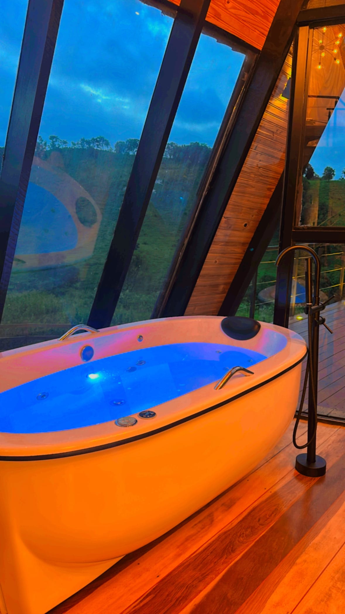 A modern private jacuzzi is situated beside floor-to-ceiling windows, offering views of the natural surroundings. The tub is softly illuminated with ambient lights, highlighting its sleek design. Wooden flooring complements the contemporary architecture, creating a serene space for relaxation.