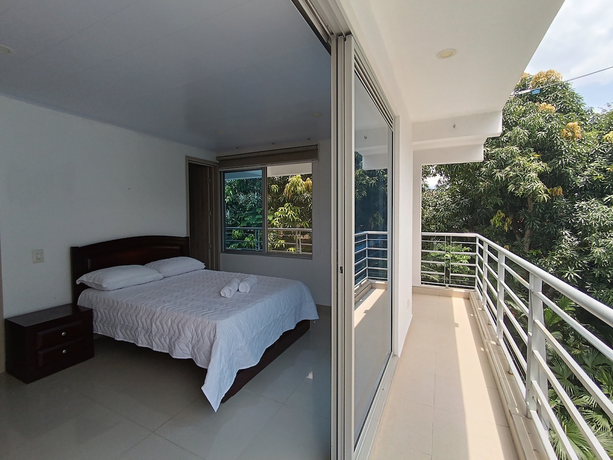 A bright bedroom features a spacious bed with fresh linens, positioned near large glass doors that open to a balcony. Natural light floods the room, while views of greenery are visible through the windows. A nightstand is placed beside the bed.