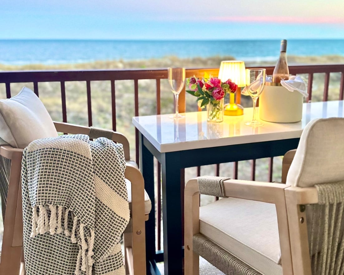 A private balcony is furnished with two cushioned chairs and a white table, adorned with a vase of flowers, a bottle of champagne, and two glasses. The soft ocean view and pastel sunset create a serene backdrop for relaxation.