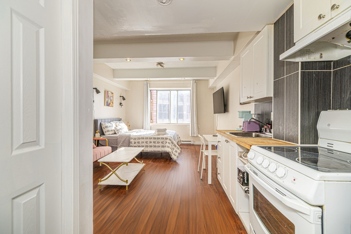A bright studio apartment features an open layout with a fully equipped kitchen to one side. A spacious living area with natural light streaming through large windows is visible, alongside a cozy sleeping area with a queen size bed. A small dining table for two is included.