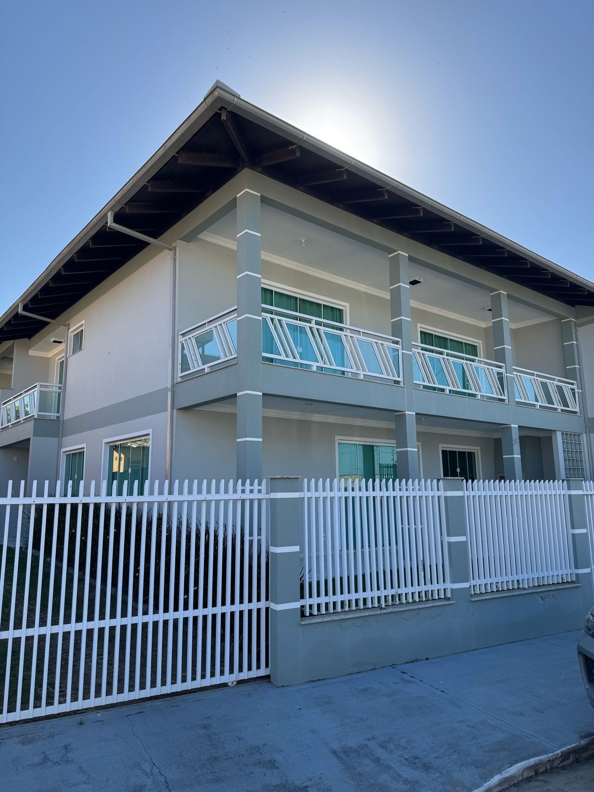 The two-story house features a light-colored exterior with white railings on the balconies. A white picket fence lines the front, providing privacy. Large windows are seen throughout, allowing natural light to fill the space. The roof has a gentle slope, enhancing its architectural profile.