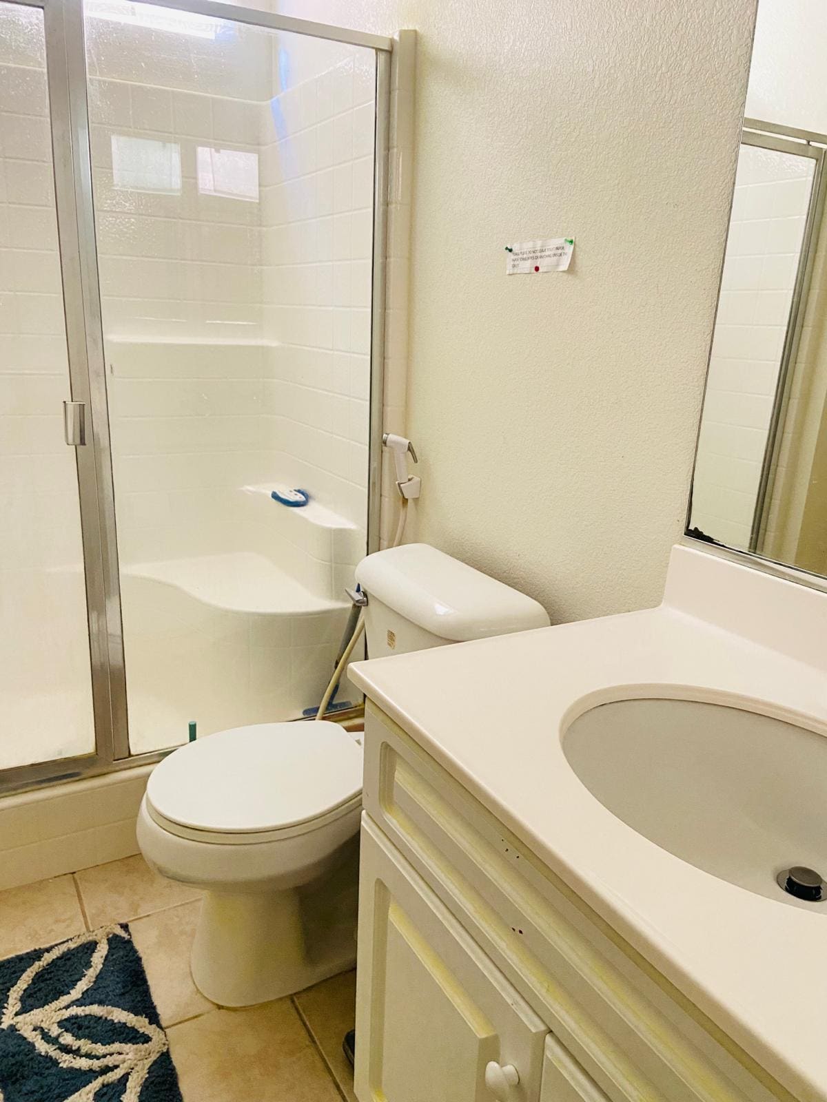 The bathroom features a walk-in shower enclosed with clear glass doors, complemented by a modern toilet and a simple white vanity with a round sink. Natural light is provided by a nearby window, and a decorative bath mat adds a subtle touch to the tile floor.