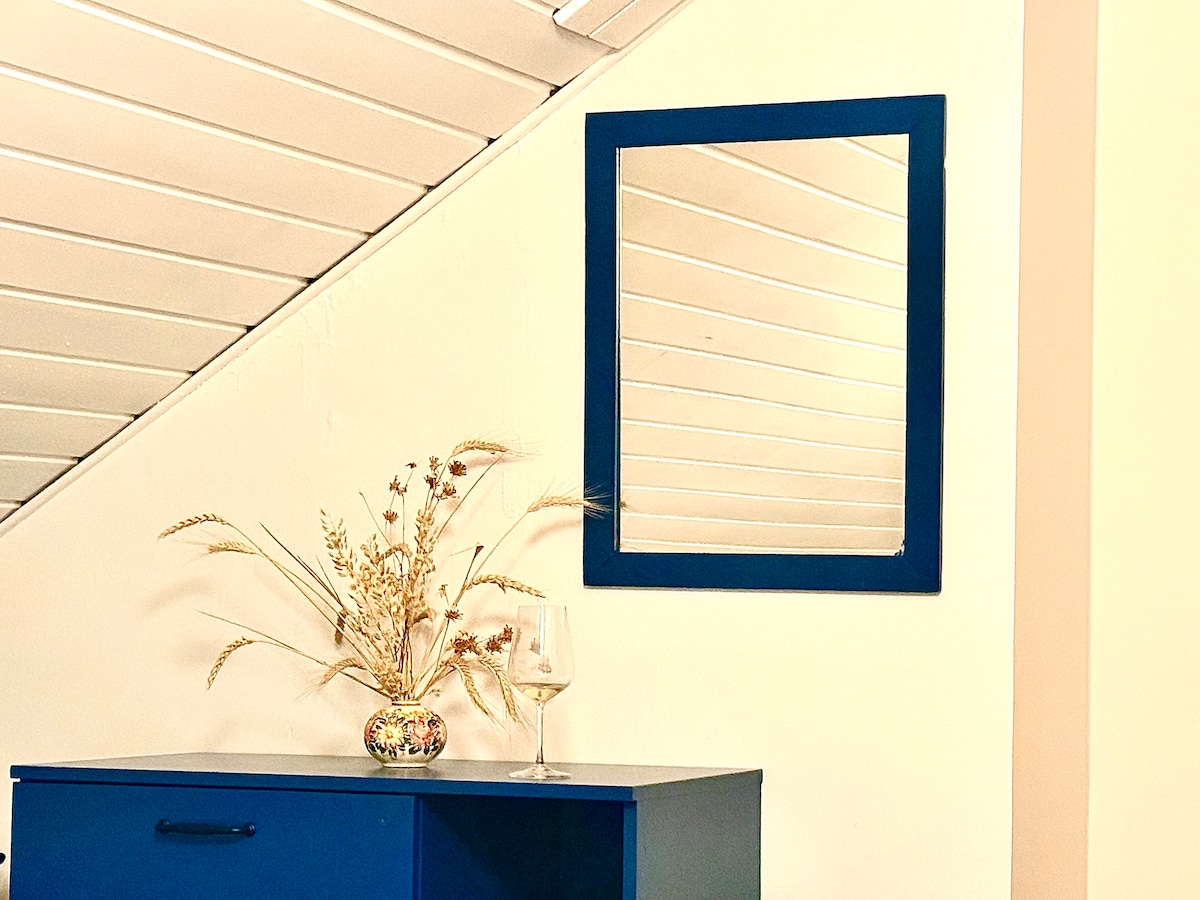 A simple blue-framed mirror is mounted on a light-toned wall, reflecting the surrounding space. A decorative arrangement of dried flowers in a small vase rests on a matching blue cabinet beneath the mirror. A clear glass sits nearby, highlighting a sense of comfort.