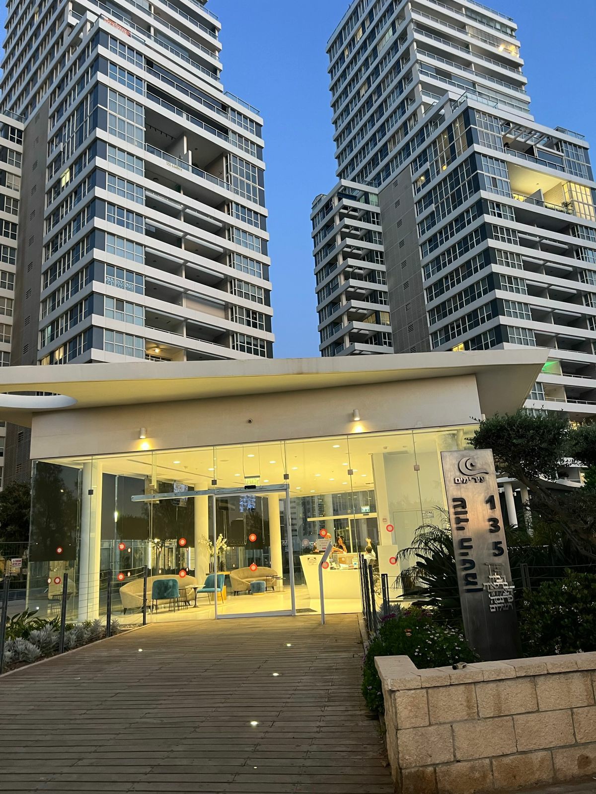 The entrance of a modern building complex is showcased, featuring sleek glass walls and bright lighting. Surrounding landscaping includes various plants, while welcoming pathways guide visitors into the lobby area. The contemporary architecture is displayed through tall structures with numerous windows.