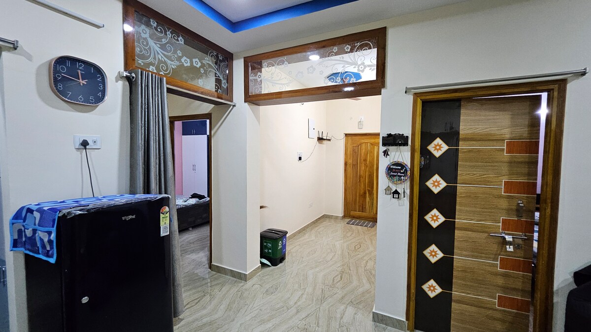 An entryway is seen featuring a wooden main door adorned with decorative elements. A clock is mounted on the wall beside a refrigerator and curtains that frame a living area visible in the background. The tiled floor adds a modern touch.