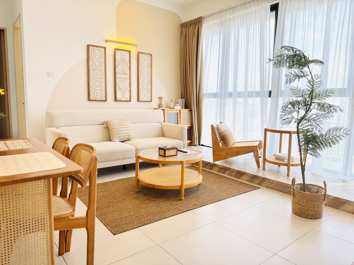 A light-filled living area features a cozy, light-colored sofa complemented by wooden furniture, including a round coffee table and a side chair. Floor-to-ceiling windows allow for natural light to enhance the ambiance, while a potted plant adds a touch of greenery.